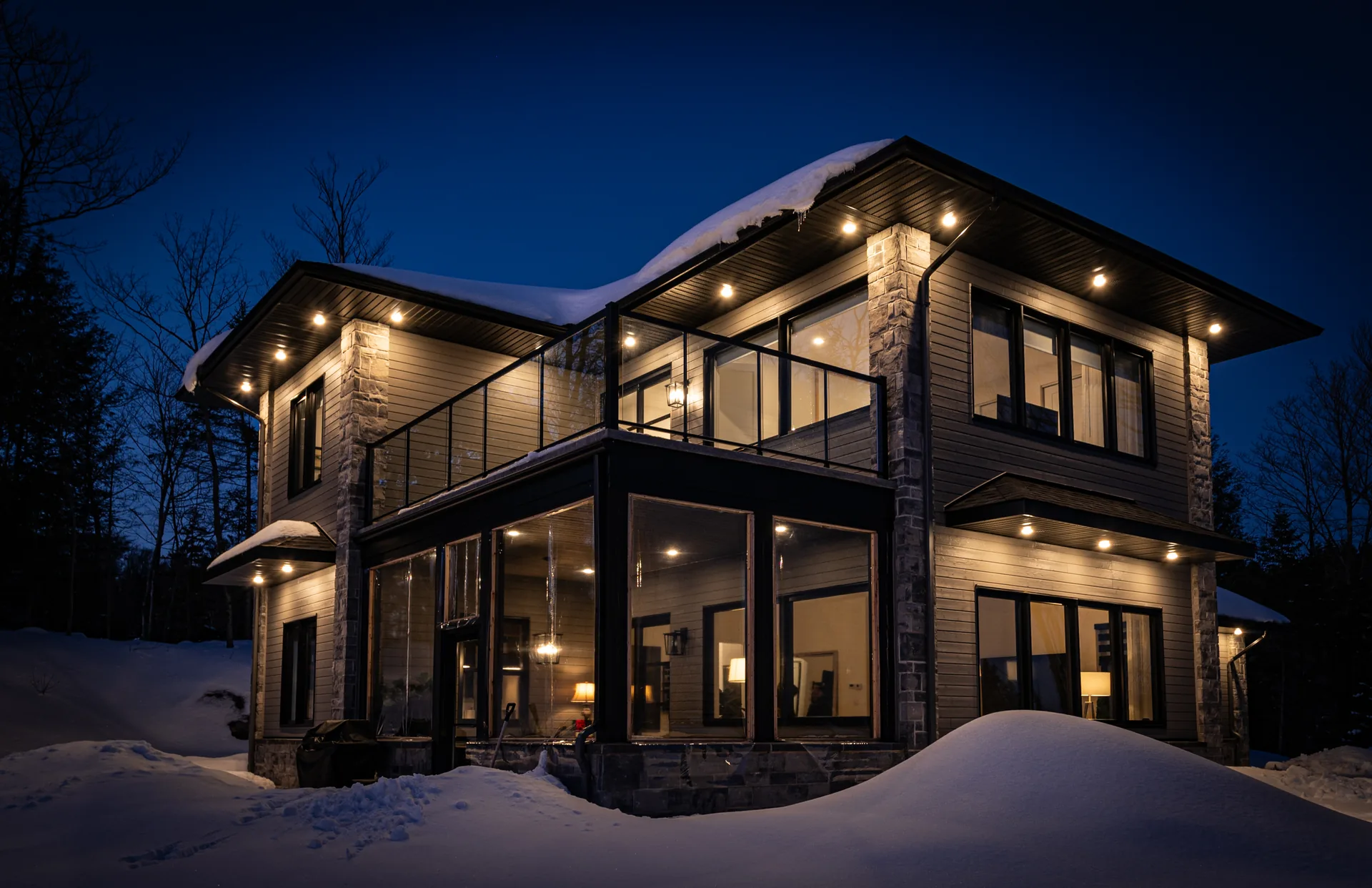 Chalet à Val-David un soir d'hiver : toit enneigé, colonnes de pierre, balcon vitré et intérieur éclairé
