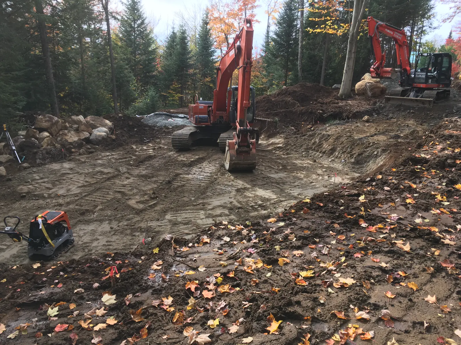 Emplacement de maison en cours de préparation à l'automne : deux pelles mécaniques dégagent et nivellent la plateforme, trépied de relevé et piquets d'implantation en place, blocs de pierre repoussés en bordure du boisé