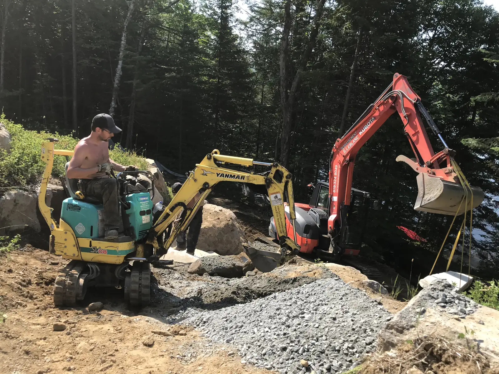 Deux pelles mécaniques travaillant à flanc de talus boisé, l'une manœuvrée par un opérateur, entre blocs de pierre et lit de gravier